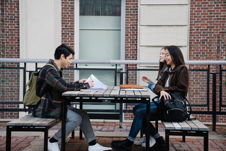 Three young adults sit at a wooden table outdoors, studying and interacting on a university campus.