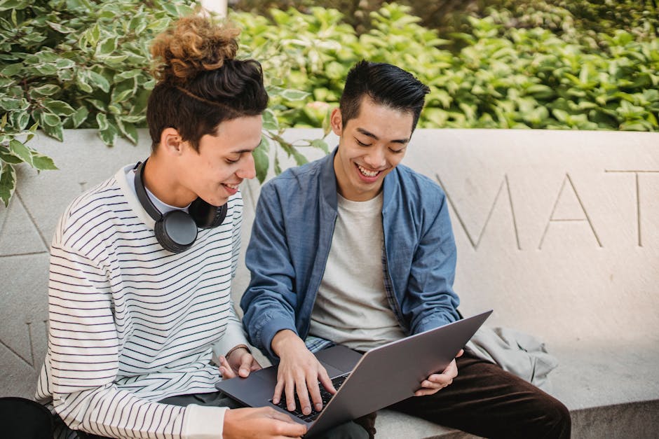 Happy young multiracial male friends in casual clothes browsing modern netbook while sitting on stone bench in verdant sunny park